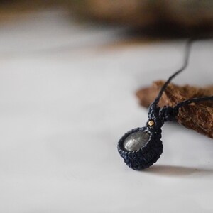 May include: A dark gray stone pendant necklace with a black cord. The pendant is wrapped in black macrame cord and has a small gold bead at the top.