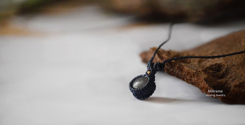 May include: A dark gray stone pendant necklace with a black cord. The pendant is wrapped in black macrame cord and has a small gold bead at the top.