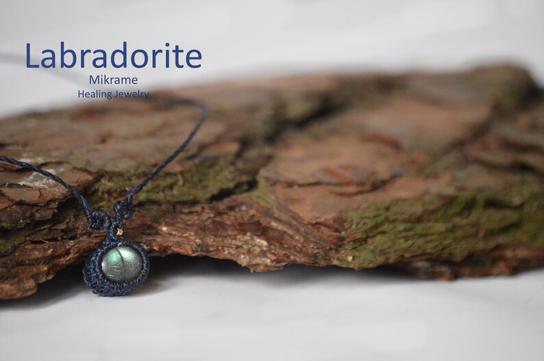 May include: A dark blue macrame necklace with a labradorite gemstone pendant. The necklace is displayed on a piece of tree bark.