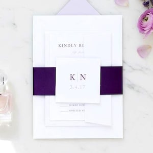 May include: A flat lay featuring wedding stationery with a purple accent. The invitation suite includes white cards, a purple belly band, and a lavender envelope. A small perfume bottle and purple flowers complete the arrangement.
