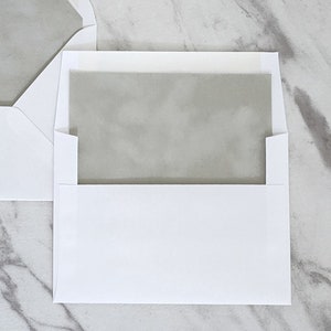 May include: A white envelope with a gray inner liner. The envelope is closed and lying flat on a marble surface.