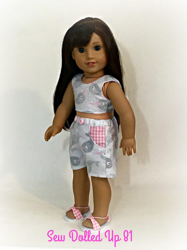 May include: A doll wearing a gray and pink outfit with a narwhal print. The outfit includes a sleeveless top and shorts. The doll is wearing pink sandals with white straps. The text "Sew Dolled Up 81" is visible at the bottom of the image.