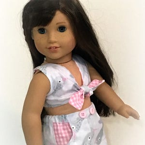 May include: A doll wearing a white and pink outfit with a narwhal print. The top is a sleeveless crop top with a pink and white gingham bow. The skirt is a high-waisted skirt with a pink and white gingham pocket. The doll has long brown hair and blue eyes. The text "Sew Dolled Up 81" is visible at the bottom of the image.