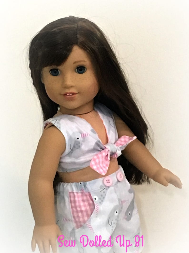 May include: A doll wearing a white and pink outfit with a narwhal print. The top is a sleeveless crop top with a pink and white gingham bow. The skirt is a high-waisted skirt with a pink and white gingham pocket. The doll has long brown hair and blue eyes. The text "Sew Dolled Up 81" is visible at the bottom of the image.