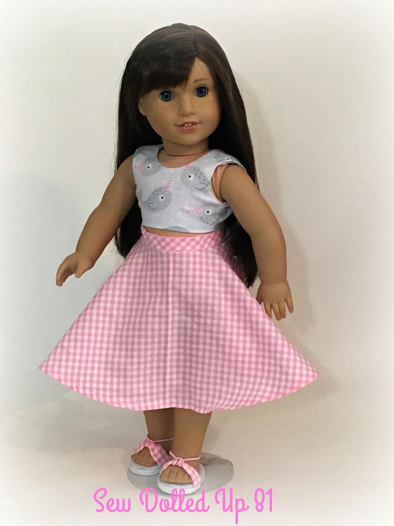 May include: A doll wearing a pink and white gingham skirt and a sleeveless top with a narwhal print. The doll has brown hair and blue eyes. The text "Sew Dolled Up 81" is at the bottom of the image.