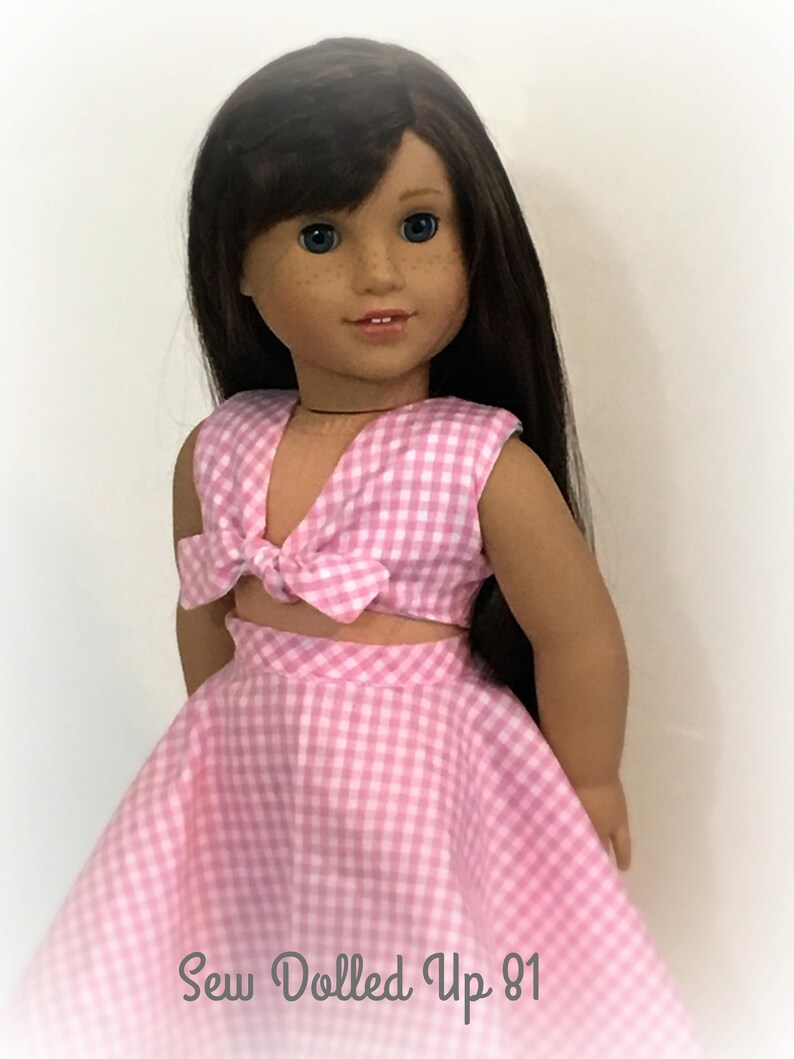 May include: A pink and white gingham doll dress with a matching top. The top has a bow tie detail. The doll is wearing the dress and has long brown hair. The text "Sew Dolled Up 81" is at the bottom of the image.