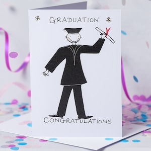May include: A white card with a black and white illustration of a person in a graduation cap and gown holding a diploma. The text "GRADUATION" is written in gold lettering at the top of the card. The text "CONGRATULATIONS" is written at the bottom of the card.