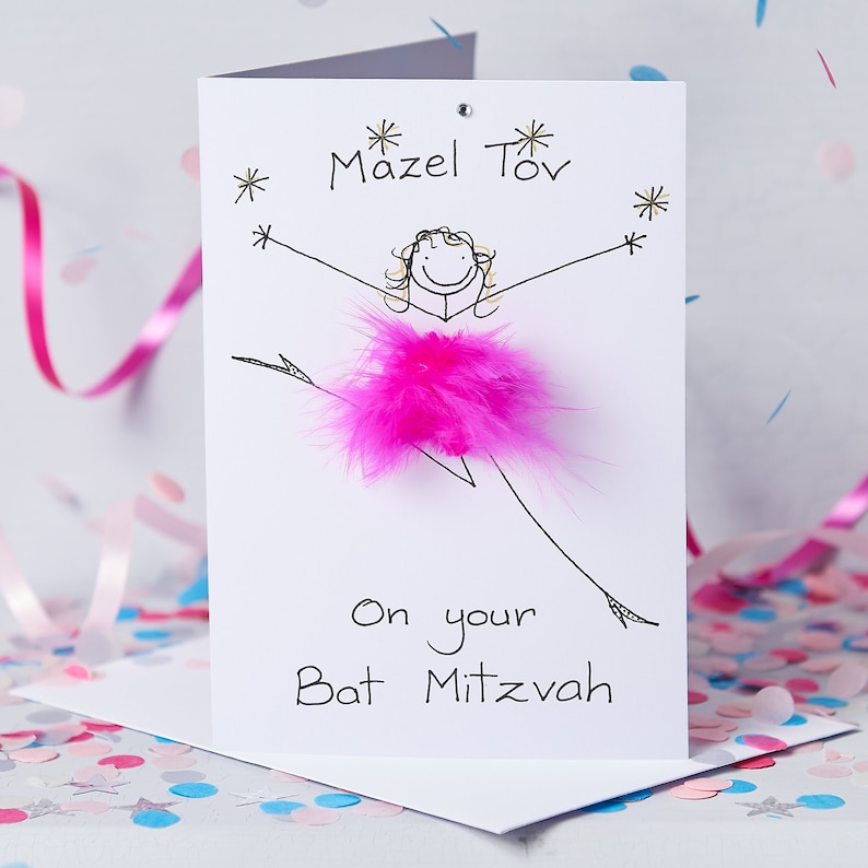 May include: A white card with the words "Mazel Tov" written in gold lettering. A drawing of a girl wearing a pink feather boa is on the card. The words "On your Bat Mitzvah" are written below the drawing.