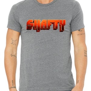 May include: A heather grey t-shirt with the word "SHAFTY" printed in orange 3D letters.