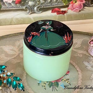 May include: A vintage green glass jar with a black lid featuring a painted illustration of a woman in a green dress. The jar is displayed with a jeweled brooch and a small decorative box, suggesting a vanity or boudoir theme.