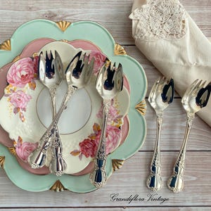 May include: A collection of vintage serving forks and plates. The plates are stacked, featuring a floral design on a white background, with a pink and mint green border. The forks are silver with ornate handles. A lace-trimmed napkin is on the right.