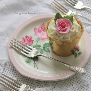 May include: A cupcake with white frosting and a pink rose on a floral plate with a pink border. Silver forks and a teacup with tea are also present. The plate has a floral design with pink roses and green leaves. The teacup is light blue with gold trim.