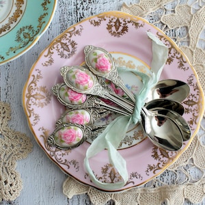 May include: A set of vintage teaspoons with rose-patterned ceramic inserts, tied with a pale green ribbon, on a pink plate with gold detailing. Another plate, in teal with gold trim, is in the background. The scene is set on a white wooden surface.
