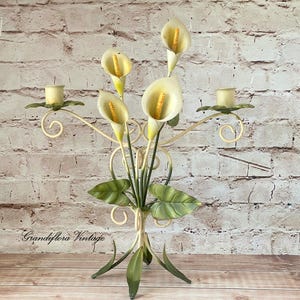 May include: A metal candelabra featuring two candle holders and a central bouquet of three cream-coloured calla lily flowers with yellow spadices. The structure includes green leaves and curved metal stems, with the text Grandiflora Vintage visible.