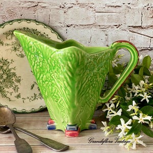 Puede incluir: Jarra de cerámica verde vintage con diseño de hojas texturizadas, asa roja y verde, y base con detalles rojos y azules. Un plato floral y cubiertos en el fondo. La jarra mide aproximadamente 20 cm de altura.