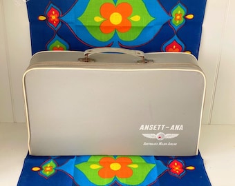 Pode incluir: Uma mala cinza vintage com acabamento branco e alça. A mala tem o texto "ANSETT-ANA Australia's Major Airline" impresso na frente. Descansa sobre um tecido azul estampado.