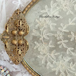 May include: Vintage gold-toned metal compact with ornate detailing and a floral lace design. The compact features a decorative clasp and a delicate embroidered floral accent. The text "Grandiflora Vintage" is visible on the lace.