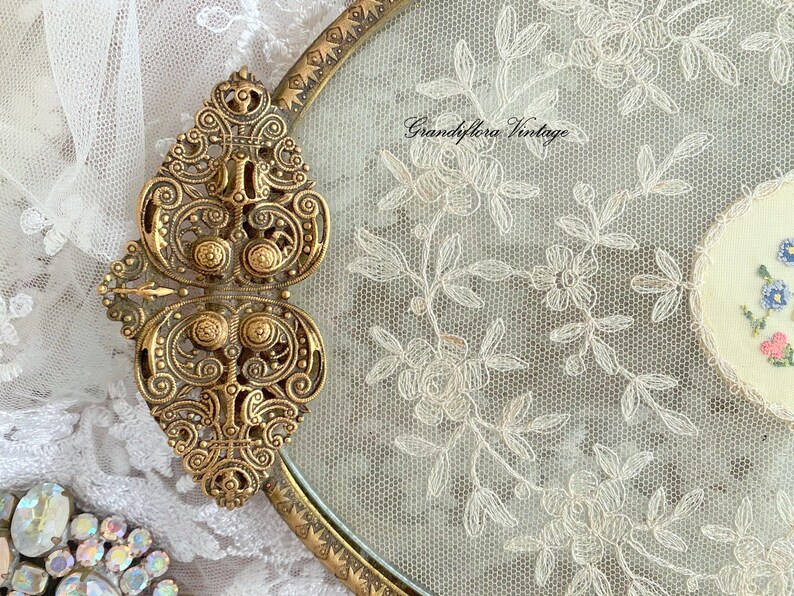 May include: Vintage gold-toned metal compact with ornate detailing and a floral lace design. The compact features a decorative clasp and a delicate embroidered floral accent. The text "Grandiflora Vintage" is visible on the lace.