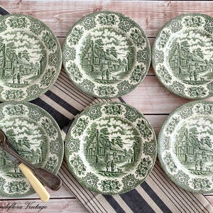 May include: Six vintage green and white plates featuring a village scene. The plates are arranged on a striped cloth, with a fork and knife. Each plate has a scalloped edge and a detailed landscape design.