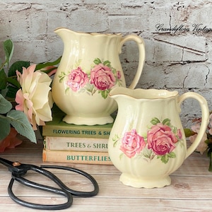 以下が含まれることがあります： ピンクのバラの花柄が描かれた、クリーム色のヴィンテージセラミックピッチャー2つ。ピッチャーは、「Flowering Trees & Shrubs」、「Trees & Shrubs」、「Butterflies」というタイトルの書籍の積み重ねの上に置かれています。