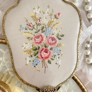 May include: An antique hand mirror with a gold-toned frame and a floral embroidered design. The embroidery features pink roses, blue forget-me-nots, and white flowers on a light beige background. The handle has an ornate, decorative design.