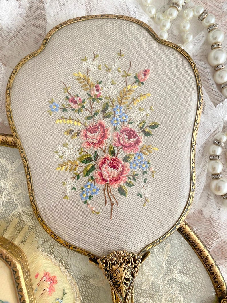 May include: An antique hand mirror with a gold-toned frame and a floral embroidered design. The embroidery features pink roses, blue forget-me-nots, and white flowers on a light beige background. The handle has an ornate, decorative design.