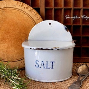 May include: White enamel salt container with a hinged lid and the word "SALT" in blue lettering. The container has a rustic, vintage appearance with visible wear and tear. A wooden breadboard and vintage silverware are in the background.