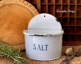 May include: White enamel salt container with a hinged lid and the word "SALT" in blue lettering. The container has a rustic, vintage appearance with visible wear and tear. A wooden breadboard and vintage silverware are in the background.
