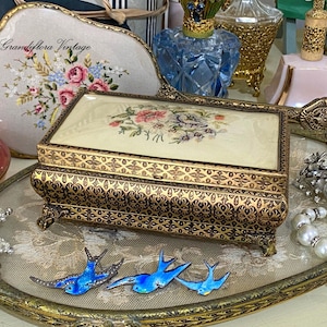 May include: A vintage jewellery box with a floral embroidered lid and ornate gold-toned metalwork. The box is rectangular with decorative feet, and is surrounded by other vintage items, including a decorative tray and blue bird brooches.