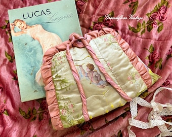 Könnte beinhalten: Vintage Lingerie-Buch mit dem Titel "LUCAS Lingerie" neben einer Satin-Handtasche mit einer Cherub-Illustration. Die Handtasche hat rosa Bandgriffe und -besatz. Der Hintergrund ist ein rosa Blumengewebe.