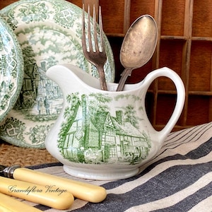 May include: A white ceramic creamer with a green and white rural scene, containing a fork and spoon. A matching plate is in the background. The creamer is on a striped cloth, with a knife and the text "Grandiflora Vintage".