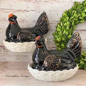 May include: Two black ceramic hen figurines with gold accents and white speckles, resting in white woven baskets. The hens have orange combs and beaks. The figurines are vintage-style decorative items.