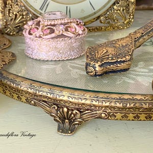 May include: An antique vanity set with a gold-toned, ornate tray, a small round trinket box, and a decorative hand mirror. The tray has a glass surface and is embellished with intricate floral and geometric patterns. The set evokes a vintage aesthetic.