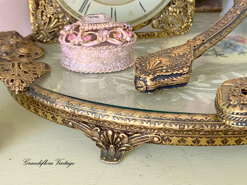 May include: An antique vanity set with a gold-toned, ornate tray, a small round trinket box, and a decorative hand mirror. The tray has a glass surface and is embellished with intricate floral and geometric patterns. The set evokes a vintage aesthetic.