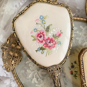 May include: An antique hand mirror with a cream-colored embroidered floral design. The mirror has a gold-toned ornate frame and handle. The embroidery features pink, red, blue, and green thread. Other vintage vanity items are in the background.