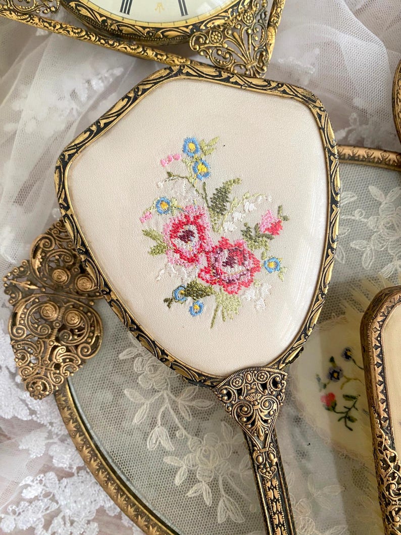 May include: An antique hand mirror with a cream-colored embroidered floral design. The mirror has a gold-toned ornate frame and handle. The embroidery features pink, red, blue, and green thread. Other vintage vanity items are in the background.