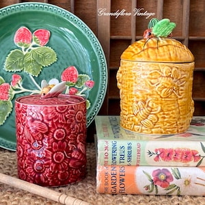 May include: A vintage still life featuring a green strawberry-themed plate, a red floral jar with a bee lid, and a yellow beehive-shaped jar with a bee lid. The jars are placed on top of vintage books. The text "Grandiflora Vintage" is visible.