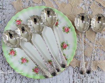 May include: Five silver-toned spoons with ornate embossed handles featuring intricate floral and scroll patterns, arranged on a white ceramic plate with a scalloped light green rim and pink rose motifs. The text Vintage Table Treasure is visible.