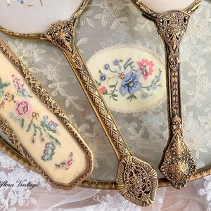 May include: Vintage vanity set featuring a hand mirror, a brush, and a small oval mirror. The set has ornate gold-toned metal frames and handles. Each piece has a cream-colored background with embroidered floral designs in pink, blue, and green.