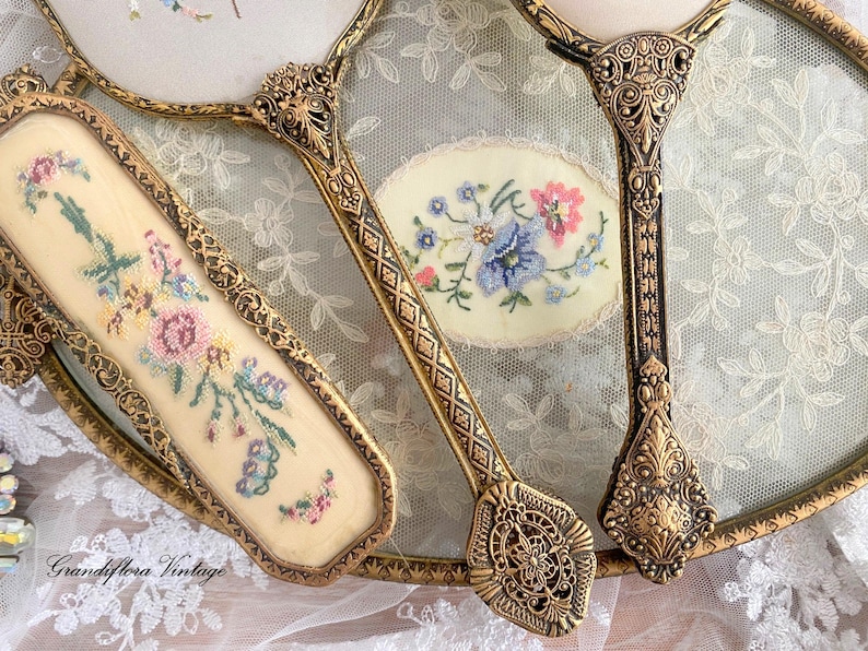 May include: Vintage vanity set featuring a hand mirror, a brush, and a small oval mirror. The set has ornate gold-toned metal frames and handles. Each piece has a cream-colored background with embroidered floral designs in pink, blue, and green.