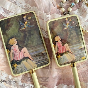Puede incluir: Dos espejos de mano vintage con mangos blancos y un marco de color crema. Los espejos presentan una ilustración caprichosa de una niña sentada en una roca junto a un lago, mirando hacia arriba a una hada volando sobre un pájaro.