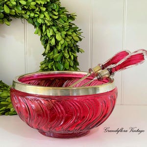 Puede incluir: Un cuenco de cristal rojo rubí con borde plateado y diseño ondulado texturizado. Dos utensilios de servir con mangos de cristal rojo en su interior. Una pieza vintage, ideal para servir fruta o aperitivos.