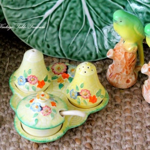 Puede incluir: Un juego de salero y pimentero vintage de cerámica amarilla con un plato y una cuchara a juego, decorado con diseños florales. También hay dos figuras de pájaros. El texto "Vintage Table Treasure" es visible.