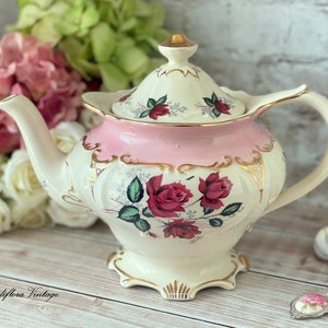 May include: A vintage teapot with a pink and white floral design. The teapot has a gold trim and a lid with a floral design. The teapot is sitting on a wooden surface.