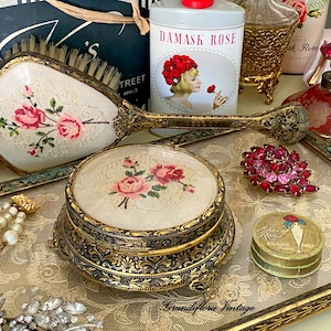 以下が含まれることがあります： 花柄のニードルポイントの蓋が付いた丸いパウダーケース、装飾的なゴールドトーンのハンドルの手鏡、赤いラインストーンのブローチ、小さなブルジョワの丸い缶、ダマスクローズのパウダー容器など、化粧台の小物のコレクション。