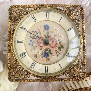 May include: A square, antique gold-toned clock with a floral embroidered face. The clock face features Roman numerals and delicate hands. The clock is encased in an ornate, filigree-style frame.