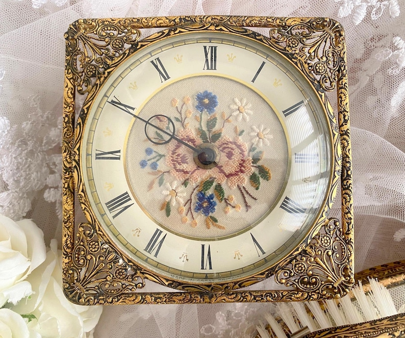 May include: A square, antique gold-toned clock with a floral embroidered face. The clock face features Roman numerals and delicate hands. The clock is encased in an ornate, filigree-style frame.