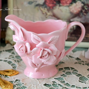 Puede incluir: Una jarra de crema de porcelana rosa con un diseño de rosas. La jarra tiene un asa y un pico. Está decorada con rosas y hojas tridimensionales.