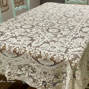May include: A rectangular off-white lace tablecloth featuring an intricate floral pattern with openwork detailing. The fabric drapes over the edges of a table, displaying a complex, repeating botanical design across the entire surface area.