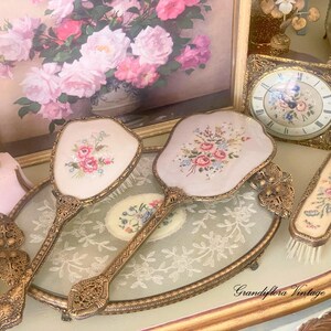 May include: Vintage vanity set featuring two hand mirrors and a brush with floral embroidery on a pale pink background. The set has ornate gold-colored metal handles and frames. A framed floral painting and a decorative clock are also visible.